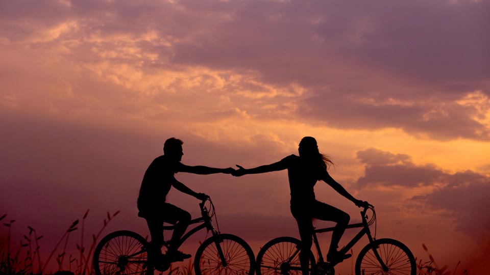 Two people in silhouette reach toward each other while riding bikes during the sunset. Photo: Unsplash