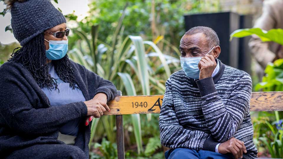 Two people wearing medical masks sit on a park bench. Photo: Unsplash