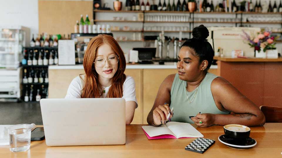 Two women look at a laptop in a coffee shop. Photo: Unsplash